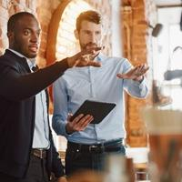 Two men talking in a cafe setting, one holding an iPad, each with an arm raised pointing across the room