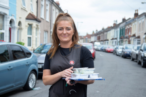 A female Bristol City Council community meals carrying some meals down a street with cars parked along the road