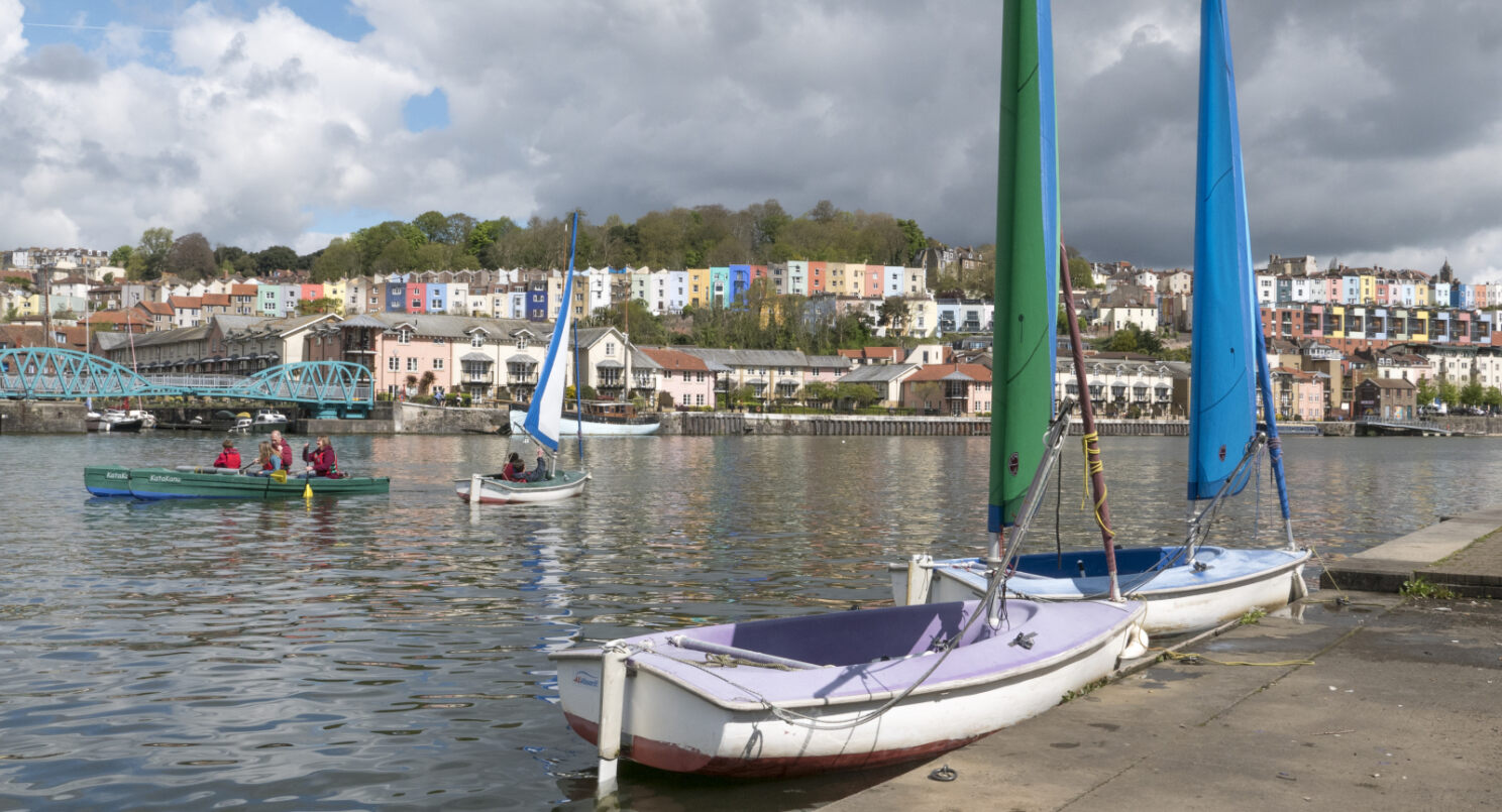 Some boats on the water, one with a green sail, one with a blue sail. More boats, a blue bridge and some colourful buildings in the background