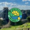 A photograph of the Clifton suspension bridge with the circular logo for the Bristol Ring city wide walking route overlaid. The logo includes a green graphic of the suspension bridge, blue water under the bridge, a yellow sun over the bridge and pale green clouds with a yellow house and a blue house.