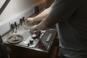 Person operating a reel-to-reel tape recorder on a workbench, adjusting controls and tape reels.