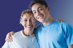 An older female and younger male standing side by side in an embrace, against a plain blue background.