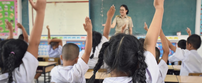 A Classroom full of children raising their hand to answer a question from the teacher.